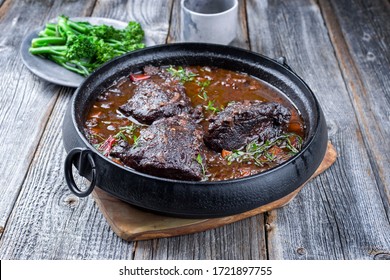 Traditional German braised beef cheeks in brown red wine sauce with carrots and broccoli offered as closeup in a modern design stewpot on an old rustic board 
