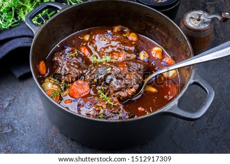 Traditional German braised beef cheeks in brown red wine sauce with carrots and onions offered as closeup in a cast iron Dutch oven on an old rustic board 