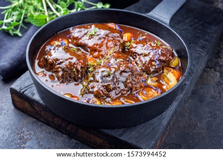 Traditional German braised beef cheeks in brown red wine sauce with carrots and onions offered as closeup in a cast iron Dutch oven on an old rustic cutting board 
