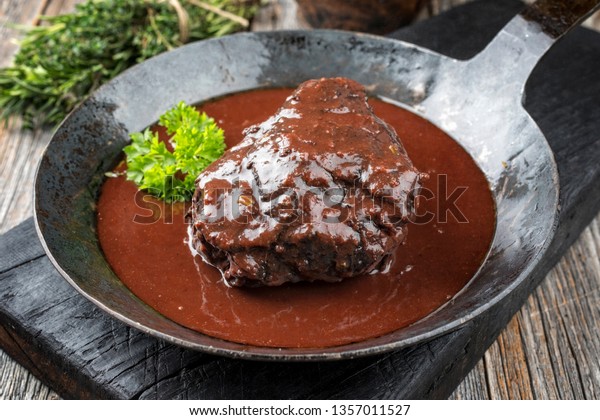 Traditional German braised beef cheeks in brown red\
wine sauce with herbs as closeup in a wrought-iron skillet on an\
old burnt board