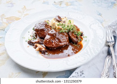 Traditional German braised pork cheeks in brown red wine sauce with mushroom and mashed potatoes as closeup on a classic design white plate 