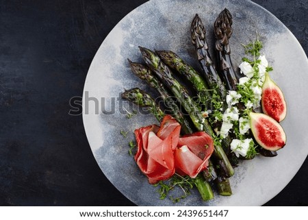 Traditional glazed purple asparagus with prosciutto ham, feta cheese and figs served as top view on a design plate with copy space left