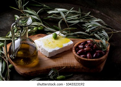 Traditional Greek Mezze Olives And Feta Decorated With Branches Of Olive Tree Served With A Jar Of Olive Oil On An Olive Boad Over A Rustic Metal Background