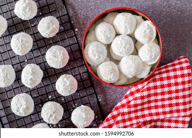 Traditional Holiday Snowball Cookies In Cookie Gift Tin With Red And White Checkered Napkin And Additional Cookies On Wire Rack