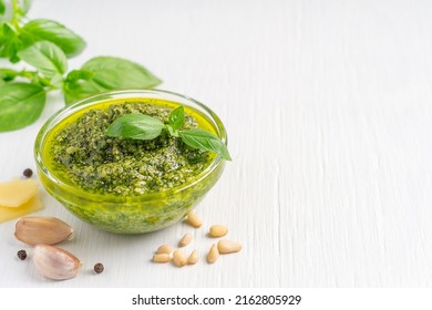 Traditional Homemade Herbal Italian Pesto Sauce Made Of Blended Green Basil Leaves, Pine Nuts, Garlic, Parmesan Cheese And Olive Oil Served In Glass Bowl On White Wooden Background With Copy Space