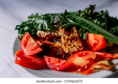 Traditional Indonesian Culinary Food. Fried Tempe In Soy Sauce, On Red And Black Pepper. Greens And Vegetables On A White Plate. Beautiful Sunlight Is Falling From The Window.