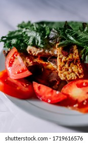 Traditional Indonesian Culinary Food. Fried Tempe In Soy Sauce, On Red And Black Pepper. Greens And Vegetables On A White Plate. Beautiful Sunlight Is Falling From The Window.