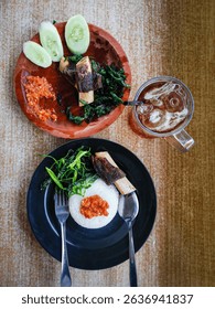 Traditional Indonesian meal with white rice, fried cassava leaves, beef ribs, sambal, and cucumber, served with iced tea on a wooden table.