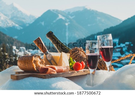 Traditional Italian food and drink outdoor in sunny winter day. Romantic alpine picnic in Dolomites with mountains background, Lambrusco cheese baguette and ham on snow.