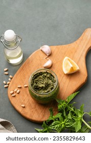 Traditional Italian pesto in a jar with green basil, pine nuts, cheese and olive oil on wooden board on a green background. A classic sauce for spaghetti or bruschetta. Top view and copy space.