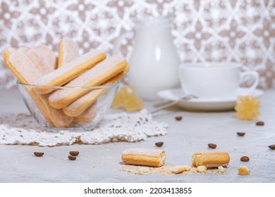 Traditional italian savoiardi or ladyfingers biscuits on a plate and pieces of cookies and sugar on the table, a white cup of coffee with a spoon and a jug of milk in the background. Selective focus