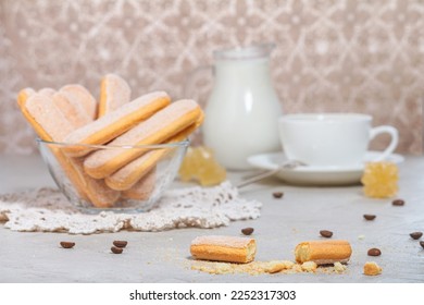 Traditional italian savoiardi or ladyfingers biscuits in a glass bowl and pieces of cookies and sugar on the table, a white cup of coffee. Selective focus