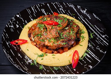 Traditional Italian Veal Osso Buco dish. Beef Ossobuco served on mashed potatoes with vegetables, Parmesan cheese, chili pepper and fresh parsley. Osso-buco dish on black plate on wooden background