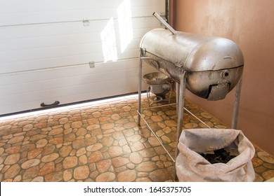 Traditional Jamaican Culture Jerk/ Grill Pan/ Drum With A White Crocus Bag Of Charcoal, And A Small One Pot Steel Metal Coal Stove On Ground Behind Near Garage Door. Enclosed Car Garage Interior. 