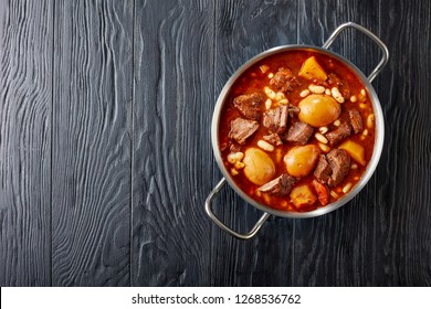 Traditional Jewish Cholent or Hamin - main dish for the Shabbat meal slow cooked beef with potato, beans and brown eggs in a metal casserole on a black wooden table, view from above, copy space