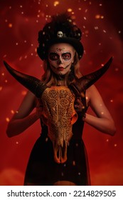 Traditional Look Of Calavera Catrina. Beautiful Sugar Skull Girl Holds An Animal Skull With Ethnic Ornaments On A Red Background With Sparks Of Fire. Day Of The Dead. Dia De Los Muertos. Halloween.