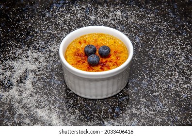 Traditional Crème Brûlée Made With Vanilla Beans And Burnt With A Blowtorch. On A Dark Surface With Scattered Sugar And Decorated With Fresh Blueberries