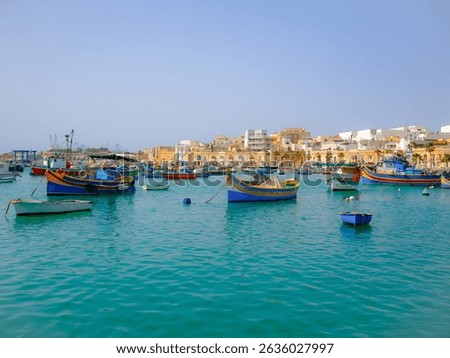 Traditional Maltese luzzus float in the turquoise waters of Marsaxlokk harbor, Malta, with a charming coastal village and clear sky creating a vibrant Mediterranean scene.