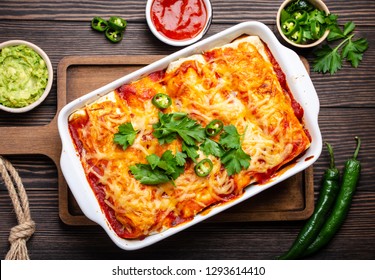 Traditional Mexican Dish Enchiladas With Meat, Chili Red Sauce And Cheese In White Casserole Dish Over Rustic Wooden Background, Served With Guacamole And Tomato Dips. Close Up, Top View