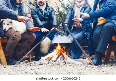 Traditional Mulled Wine With Slices Of Orange Preparing In Pot Over Burning Logs In Winter In Countryside, Friends In Winter Day