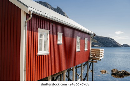 Traditional Norwegian rorbu cabins on stilts, situated by the serene waters of a fjord. These iconic red wooden cabins offer a picturesque view of the surrounding mountains and clear blue sky - Powered by Shutterstock