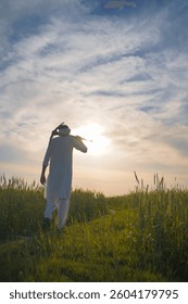 A traditional Pakistani Indian farmer walks through a lush green field at sunset, carrying a tool on his shoulder.