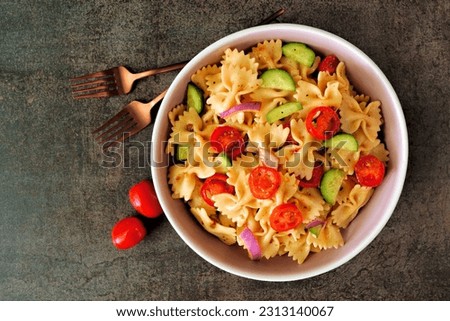Traditional pasta salad with tomatoes, onion and cucumbers. Overhead view on a dark stone background.