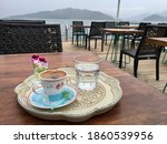 Traditional presentation of turkish coffee in ceramic cup and vintage tray with turkish delight, glass of water and pink fresh flowers. Outside garden of an empty cafe with foggy sea view. 