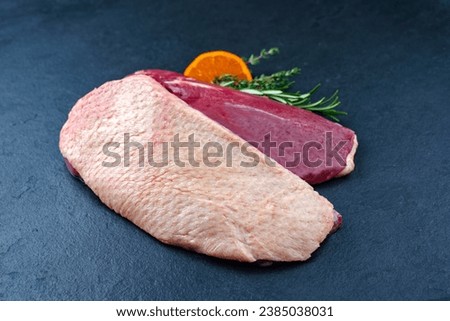 Traditional raw gourmet duck breast filet with skin offered with herbs and orange slice as close-up on a black board 