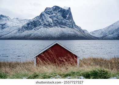 Traditional red Norwegian cabin by a tranquil fjord, framed by a dramatic snow-capped mountain in the background. A perfect depiction of serene Scandinavian nature and rustic charm - Powered by Shutterstock