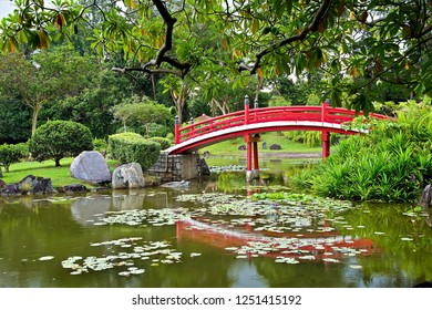 Traditional Red Wooden Bridge Over A Pond With Water Lilies Set In A Beautiful Japanese Zen Garden In Evening Light