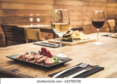 Traditional Restaurant Atmosphere With Tuna Tataki And Burger With Chips In The Background On Rustic Wooden Table And Glass Of White Wine