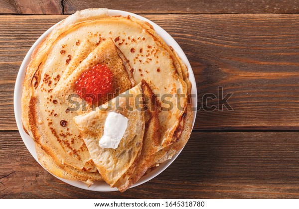 Traditional Russian Crepes Blini stacked in a plate\
with red caviar, fresh sour creamon dark wooden table. Maslenitsa\
Russian festival meal. Russian food, russian kitchen. Top view.\
Space for text