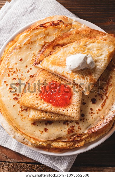 Traditional Russian Crepes Blini stacked in a plate\
with red caviar, fresh sour creamon dark wooden table. Maslenitsa\
traditional Russian festival meal. Russian food, russian kitchen.\
Close up.