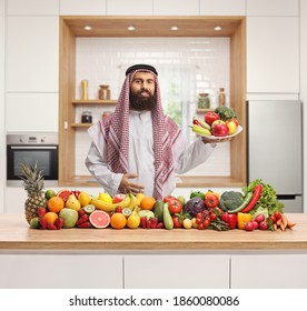 Traditional Saudi Arab Man In A Kitchen Holding A Plate Of Fruits And Vegetables And Standing Behind A Pile Of Fruits And Vegetables 