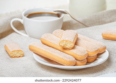 Traditional savoiardi biscuits or ladyfingers cookies on a white plate and a white cup of coffee with a spoon on a napkin. Selective focus