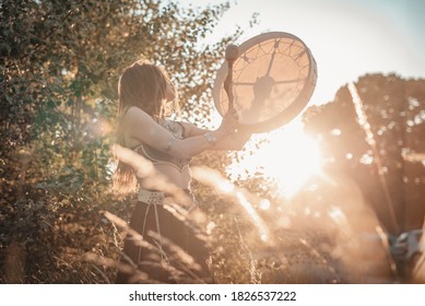 Ein traditionelles slawisches Schamanenmädchen, das die Trommel schlägt und die Naturgeister in einer herrlichen Umgebung mit Sonnenuntergang erweckt