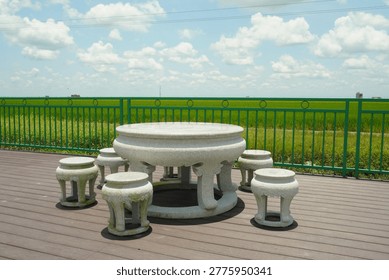 A traditional stone table and stools set on a wooden deck overlooking a lush green paddy field under a bright blue sky with fluffy white clouds.