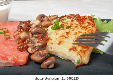 Traditional Swiss Dish Is A Sliced Potato Pancake With Pieces Of Red Salmon Fish And Fried Mushrooms On A Black Board, With White Cream Sauce In A Glass Saucepan And A Fork, Close-up.