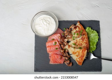 Traditional Swiss Dish Is A Sliced Potato Pancake With Pieces Of Red Salmon Fish And Fried Mushrooms On A Black Board, With White Cream Sauce In A Glass Saucepan And A Fork, Top View, Copy Space.