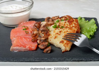 A Traditional Swiss Dish Is A Sliced Potato Pancake With Pieces Of Red Salmon Fish And Fried Mushrooms On A Black Board, With White Cream Sauce In A Glass Saucepan And A Fork.