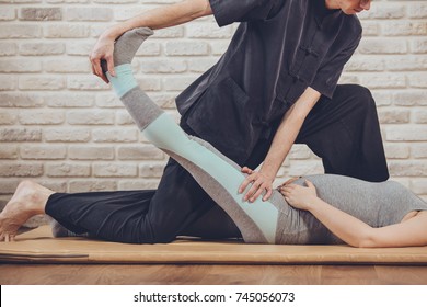 Traditional Thai Massage of pregnant woman lying on the mat in yoga studio. Young white masseur dressed in black uniform stretches her leg with his hands. Brick wall in the background. Health concept