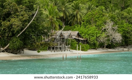 Traditional thatched roof rustic beach huts nestled amongst rainforest trees on white sandy beach overlooking turquoise ocean water on tropical island in Raja Ampat, West Papua, Indonesia