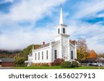 Traditional white American wooden church at the foot of a forested hill shrouded in morning fog on a autumn day