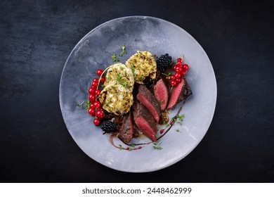 Traditionally roasted saddle of venison fillet with South Tyrolean bread dumplings, redcurrants and blackberries in game sauce served as top view on a Nordic design plate with copy space 