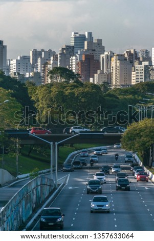 
Traffic of cars and motorcycles on an avenue passing under a viaduct, surrounded by trees and green with the buildings of the city of sao paulo in the background