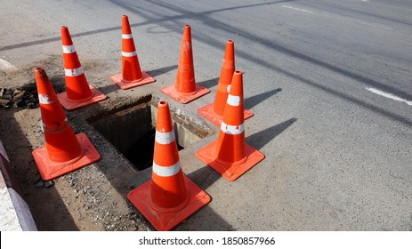 Traffic Cones Around The Manhole Open.Orange Plastic Cones On The Roadway Warn Of Potentially Dangerous Traffic Due To Sewer Repair Work. On A Paved Road Background With A Copy Area. Selective Focus