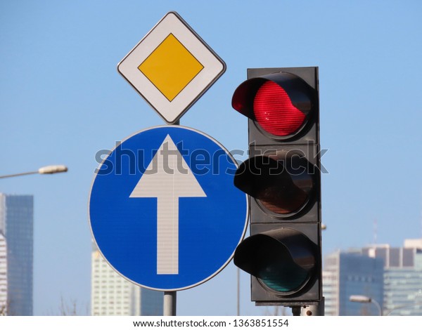 Traffic lights with\
the red signal on and road signs prohibitory Ahead only, Priority\
road, blue sky and blurred high rise buildings in the background,\
on sunny spring day