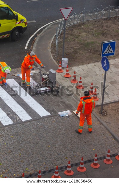 Traffic line painting. Workers are\
painting white street lines on pedestrian crossing. Road cones with\
orange and white stripes in background,\
standing