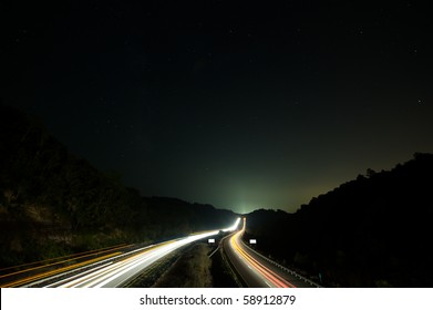 Traffic moving along interstate under starry sky toward glowing city beyond ridge.